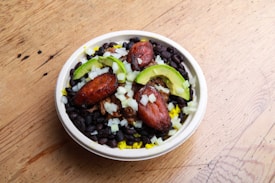 A bowl filled with black beans, sliced avocado, fried plantains, chopped onions, and what appears to be yellow rice or possibly shredded cheese. The ingredients are arranged in a visually appealing manner, with the colors contrasting against each other. The bowl is set on a wooden surface, which adds a rustic element to the presentation.