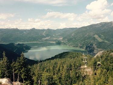 A scenic landscape featuring a large, calm lake surrounded by lush green forests and mountains under a partly cloudy sky. Cable cars are seen traversing from one side of the forest to another, providing a clear view of the beautiful natural setting.