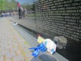 A reflective black stone wall inscribed with numerous names of individuals is viewed at an angle. Nearby, a cluster of white flowers with blue ribbons lies on the pavement. In the background, a group of people, including a child sitting on an adult's shoulders, are visible, suggesting a memorial or commemorative setting.
