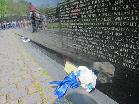 A reflective black stone wall inscribed with numerous names of individuals is viewed at an angle. Nearby, a cluster of white flowers with blue ribbons lies on the pavement. In the background, a group of people, including a child sitting on an adult's shoulders, are visible, suggesting a memorial or commemorative setting.