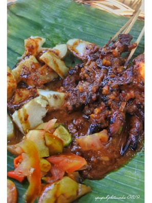 A dish is presented on a banana leaf, featuring grilled meat skewers covered in a rich, brown sauce. Accompanying the skewers are slices of rice cake, along with fresh cucumber and tomato slices, arranged on the side.