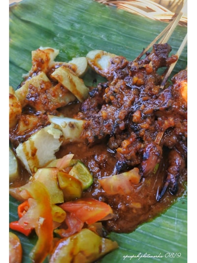 A dish is presented on a banana leaf, featuring grilled meat skewers covered in a rich, brown sauce. Accompanying the skewers are slices of rice cake, along with fresh cucumber and tomato slices, arranged on the side.