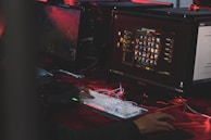 A person is sitting in front of multiple computer monitors, one displaying a video game character selection screen. The environment has low lighting with a red hue, suggesting an intense or focused atmosphere. The person is actively interacting with a mechanical keyboard with RGB lighting.