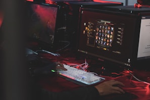 A person is sitting in front of multiple computer monitors, one displaying a video game character selection screen. The environment has low lighting with a red hue, suggesting an intense or focused atmosphere. The person is actively interacting with a mechanical keyboard with RGB lighting.