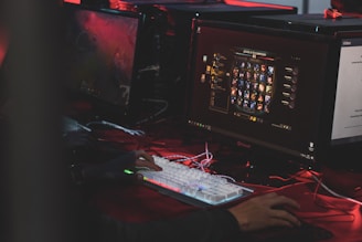 A person is sitting in front of multiple computer monitors, one displaying a video game character selection screen. The environment has low lighting with a red hue, suggesting an intense or focused atmosphere. The person is actively interacting with a mechanical keyboard with RGB lighting.