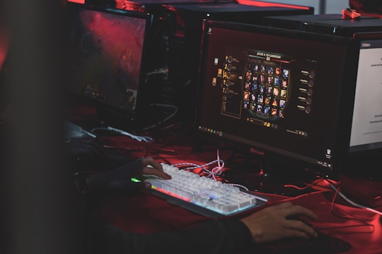 A person is sitting in front of multiple computer monitors, one displaying a video game character selection screen. The environment has low lighting with a red hue, suggesting an intense or focused atmosphere. The person is actively interacting with a mechanical keyboard with RGB lighting.