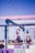 A vibrant outdoor terrace with a pastel color scheme featuring shades of pink, purple, and blue. The area includes umbrellas, glass panels, and string lights overhead, suggesting a relaxed and inviting atmosphere with a view of a distant landscape.