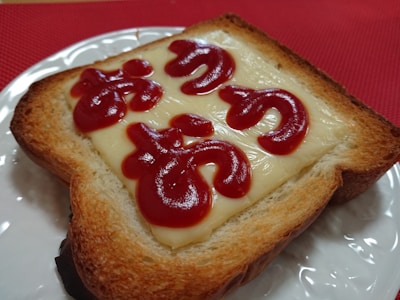 A slice of toasted bread topped with melted cheese, featuring red ketchup writing on the cheese surface. The toast is placed on a white plate with a red background underneath.