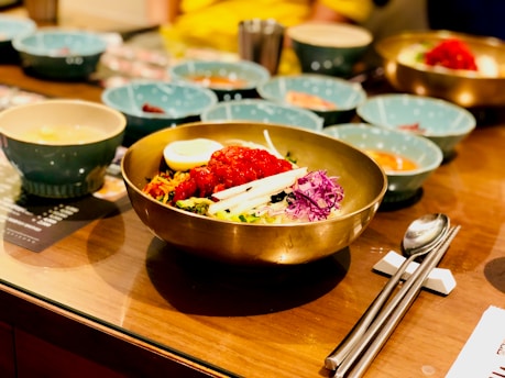 A bowl of Korean bibimbap is placed on a dining table, surrounded by several blue dishes and small bowls. The dish features vibrant ingredients such as red chili paste, sliced vegetables, shredded purple cabbage, white radish, and a halved boiled egg on top. Silver utensils, including chopsticks and a spoon, rest beside the bowl.
