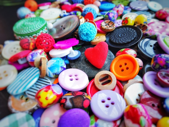 A colorful assortment of various buttons scattered across a surface, featuring a prominent red heart-shaped button in the center. The buttons vary in size, shape, and design, including round, square, and textured patterns.