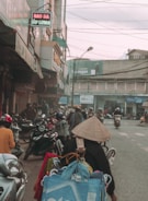 A bustling street scene featuring numerous motorbikes and pedestrians. The foreground has a person wearing a traditional conical hat, surrounded by bags and baskets, likely a street vendor. Shops line the sides of the street, some with signs in Vietnamese. Power lines crisscross above, and the atmosphere appears busy and slightly overcast.