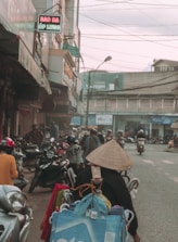 A bustling street scene featuring numerous motorbikes and pedestrians. The foreground has a person wearing a traditional conical hat, surrounded by bags and baskets, likely a street vendor. Shops line the sides of the street, some with signs in Vietnamese. Power lines crisscross above, and the atmosphere appears busy and slightly overcast.