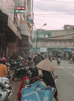 A bustling street scene featuring numerous motorbikes and pedestrians. The foreground has a person wearing a traditional conical hat, surrounded by bags and baskets, likely a street vendor. Shops line the sides of the street, some with signs in Vietnamese. Power lines crisscross above, and the atmosphere appears busy and slightly overcast.