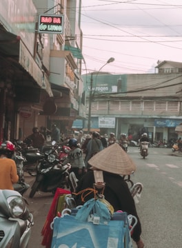 A bustling street scene featuring numerous motorbikes and pedestrians. The foreground has a person wearing a traditional conical hat, surrounded by bags and baskets, likely a street vendor. Shops line the sides of the street, some with signs in Vietnamese. Power lines crisscross above, and the atmosphere appears busy and slightly overcast.