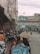 A bustling street scene featuring numerous motorbikes and pedestrians. The foreground has a person wearing a traditional conical hat, surrounded by bags and baskets, likely a street vendor. Shops line the sides of the street, some with signs in Vietnamese. Power lines crisscross above, and the atmosphere appears busy and slightly overcast.