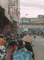 A bustling street scene featuring numerous motorbikes and pedestrians. The foreground has a person wearing a traditional conical hat, surrounded by bags and baskets, likely a street vendor. Shops line the sides of the street, some with signs in Vietnamese. Power lines crisscross above, and the atmosphere appears busy and slightly overcast.