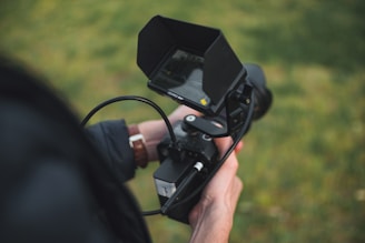 A person holds a handheld digital video camera with a flip-out screen and various controls. The camera is aimed forward and surrounded by a grassy background, partially visible in the blurred areas.