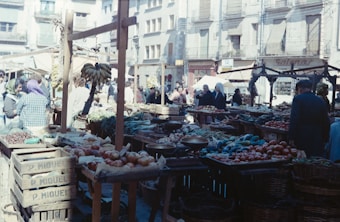 A bustling outdoor market scene with various stalls displaying fresh produce such as tomatoes, bananas, and other fruits and vegetables. People are walking around, some engaged in conversations, creating a vibrant atmosphere. The background shows old buildings, adding a rustic charm to the setting as sunlight casts shadows across the area.