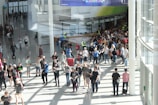 A large group of people gathered in a spacious, well-lit indoor area with glass walls. Many individuals are moving around, some in conversation and others holding bags. There is a large, visible queue in the background, organized by barriers, and several people are wearing gaming-related apparel. Signs with text hang from the ceiling.