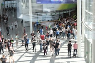 A large group of people gathered in a spacious, well-lit indoor area with glass walls. Many individuals are moving around, some in conversation and others holding bags. There is a large, visible queue in the background, organized by barriers, and several people are wearing gaming-related apparel. Signs with text hang from the ceiling.