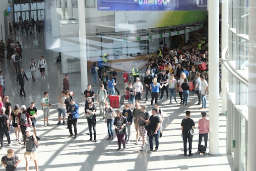 A large group of people gathered in a spacious, well-lit indoor area with glass walls. Many individuals are moving around, some in conversation and others holding bags. There is a large, visible queue in the background, organized by barriers, and several people are wearing gaming-related apparel. Signs with text hang from the ceiling.