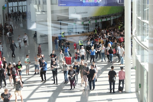 A large group of people gathered in a spacious, well-lit indoor area with glass walls. Many individuals are moving around, some in conversation and others holding bags. There is a large, visible queue in the background, organized by barriers, and several people are wearing gaming-related apparel. Signs with text hang from the ceiling.