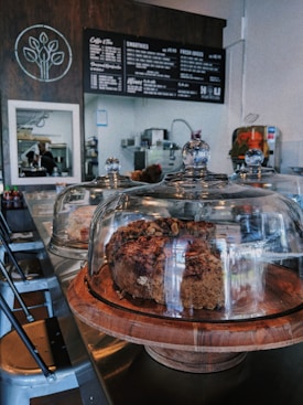 A cozy cafe setting with a modern, minimalist interior. Glass domes cover baked goods on wooden trays placed on a metal countertop. A chalkboard menu hangs on a wooden wall featuring various items including smoothies and juices. In the background, there is kitchen equipment and a barista at work. Chairs are neatly arranged along the counter.