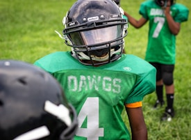 A young football player wearing a green jersey with the word 'DAWGS' and the number four on it stands on a grassy field. The player is also wearing a black helmet with a visor. Another player in the background is visible, wearing a similar green jersey with the number seven.