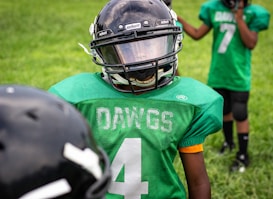 A young football player wearing a green jersey with the word 'DAWGS' and the number four on it stands on a grassy field. The player is also wearing a black helmet with a visor. Another player in the background is visible, wearing a similar green jersey with the number seven.