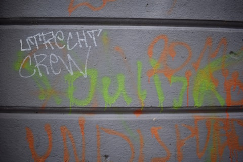 Graffiti on a gray wall featuring a combination of white, green, and orange spray-painted words. The lettering includes the phrase 'Utrecht Crew' in prominent white writing. The wall has a textured surface with horizontal lines running across.