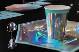 A close-up of a party table setting with a holographic paper cup and matching plate on a black surface. A shiny metal spoon with an ornate handle is placed next to the plate. The cup and plate display reflective colors, creating a festive atmosphere.
