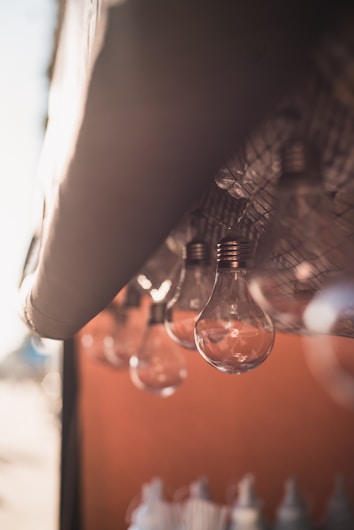 A row of clear incandescent light bulbs is suspended from a metal grid above. The background is softly blurred, with warm sunlight casting gentle reflections on the bulbs, creating an inviting and serene ambiance.