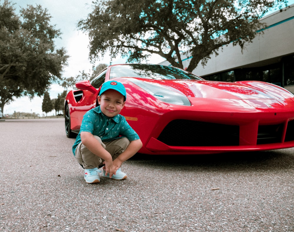 toddler in green polo shirt squatting beside red luxury car
