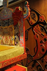 A richly decorated piece of furniture with intricate patterns and vibrant colors is placed next to a wall with abstract art. The furniture features floral and ornate designs in red, blue, and gold, while the wall art includes stylized, curved shapes in black and red.