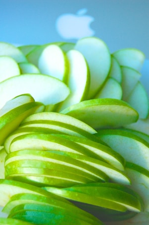Sliced green apples are arranged closely together, showcasing their fresh and crisp texture. A blurred Apple logo is visible in the background, adding a technological or branding aspect to the composition.