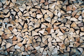 A large pile of chopped firewood pieces, neatly stacked and closely packed together. The wood displays various shades of brown, beige, and tan, highlighting the natural textures and grain patterns.