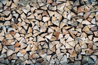 A large pile of chopped firewood pieces, neatly stacked and closely packed together. The wood displays various shades of brown, beige, and tan, highlighting the natural textures and grain patterns.