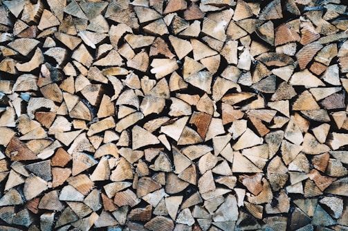 A large pile of chopped firewood pieces, neatly stacked and closely packed together. The wood displays various shades of brown, beige, and tan, highlighting the natural textures and grain patterns.