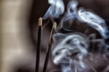 Two incense sticks are emitting swirling patterns of smoke against a dark background. The smoke is curling gracefully, creating soft, flowing shapes as it ascends.