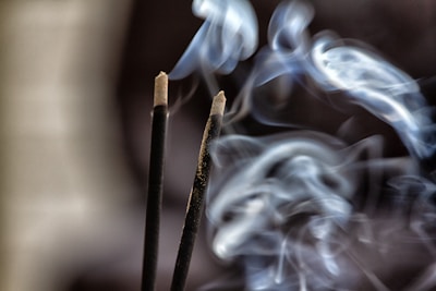 Two incense sticks are emitting swirling patterns of smoke against a dark background. The smoke is curling gracefully, creating soft, flowing shapes as it ascends.