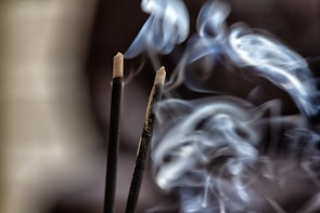 Two incense sticks are emitting swirling patterns of smoke against a dark background. The smoke is curling gracefully, creating soft, flowing shapes as it ascends.