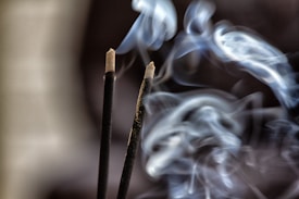 Two incense sticks are emitting swirling patterns of smoke against a dark background. The smoke is curling gracefully, creating soft, flowing shapes as it ascends.