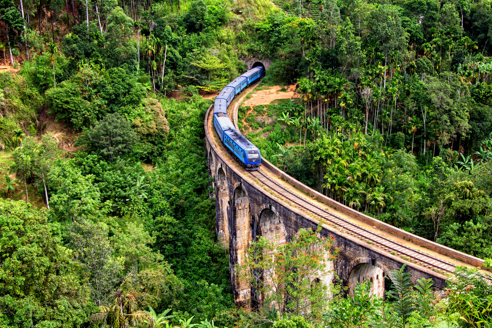 Un tren azul cruzando el puente de los Nueve Arcos entre la vegetación, cerca de Ella