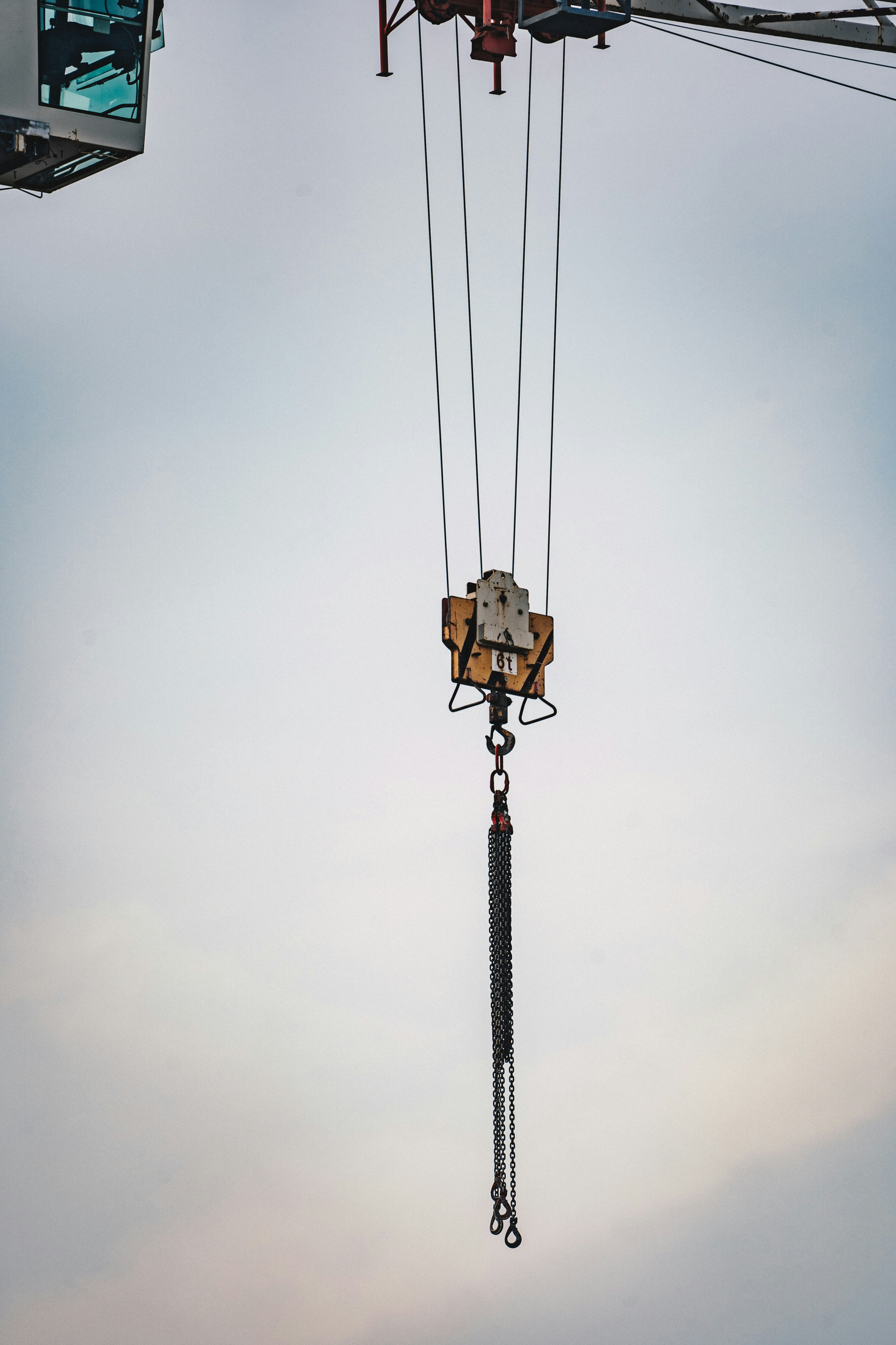 A crane's lifting mechanism is suspended mid-air with extended metal chains hanging down. The background features a soft, cloudy sky.