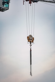 A crane's lifting mechanism is suspended mid-air with extended metal chains hanging down. The background features a soft, cloudy sky.