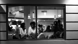 A black and white photograph taken from outside a building looking through a window into a crowded room. Several people are gathered at a table engaging in conversation. Overhead lights illuminate the scene, casting soft shadows. The atmosphere appears social and busy, with people in the background standing and mingling.