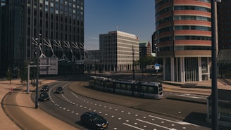 A modern urban scene featuring tall office buildings with distinctive architectural designs. A tram is moving through an intersection, and several cars are on the road. The area appears clean and well-organized, with clear skies above.