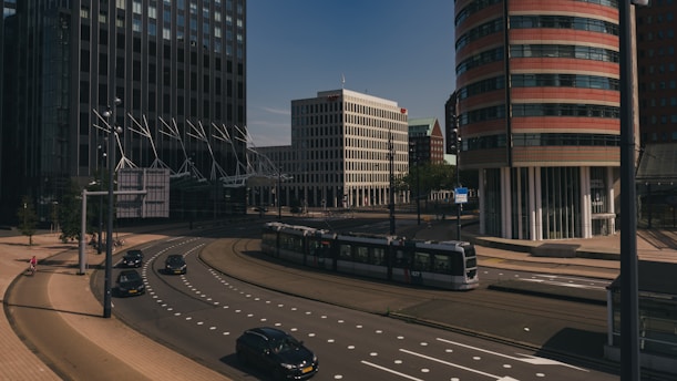 A modern urban scene featuring tall office buildings with distinctive architectural designs. A tram is moving through an intersection, and several cars are on the road. The area appears clean and well-organized, with clear skies above.