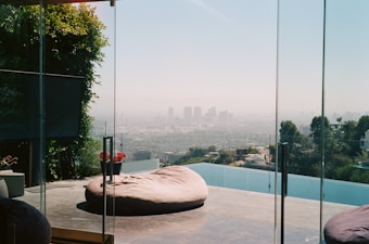 A modern, luxurious open-air living space features a large glass wall offering a panoramic view of a city skyline. A comfortable, large round outdoor cushion rests on the stone-tiled patio near an infinity pool. The scene is framed by green foliage and a few potted plants with red flowers.