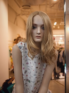 A mannequin with long blonde hair and dark eye makeup is dressed in a sleeveless white top featuring colorful patterns. It is positioned in a clothing store with a softly lit, modern interior.